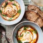 two bowls of cilbir on a light blue table with two slices of sourdough bread, a spoon and a glass of pinot gris