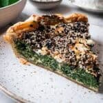 slice of spinach quiche on a white plate with spinach and goat cheese in the background