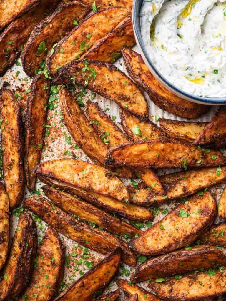 roasted Mediterranean potato wedges on a tray topped with parsley with a yogurt dip on the side.