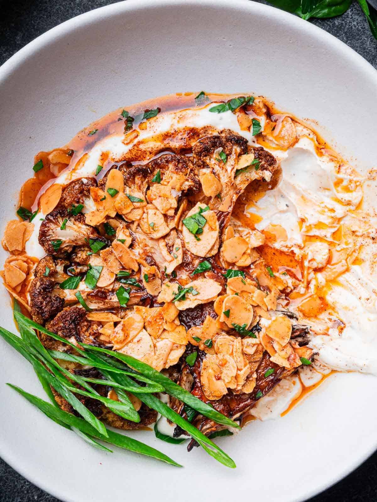 roasted cauliflower steaks on tahini yogurt sauce with almond paprika butter in a white bowl.