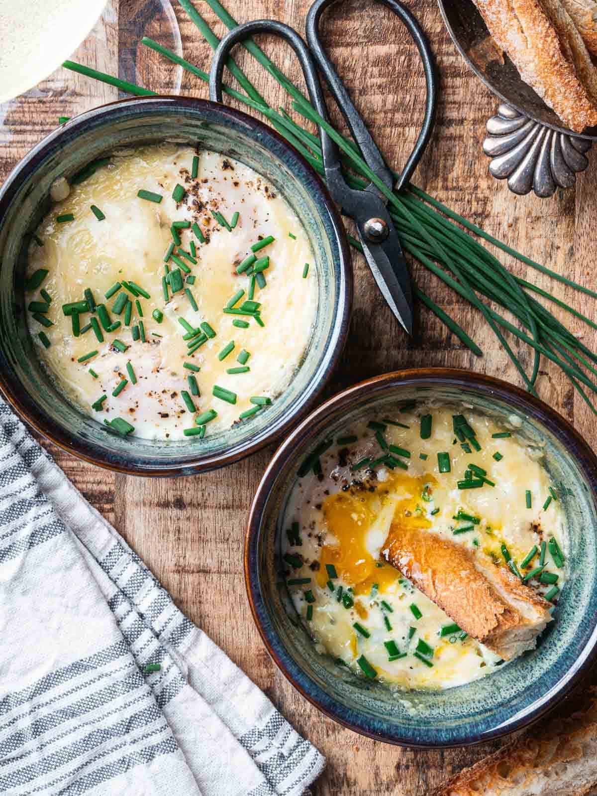 two blue ramekins with French baked eggs topped wth chives and crusty bread.