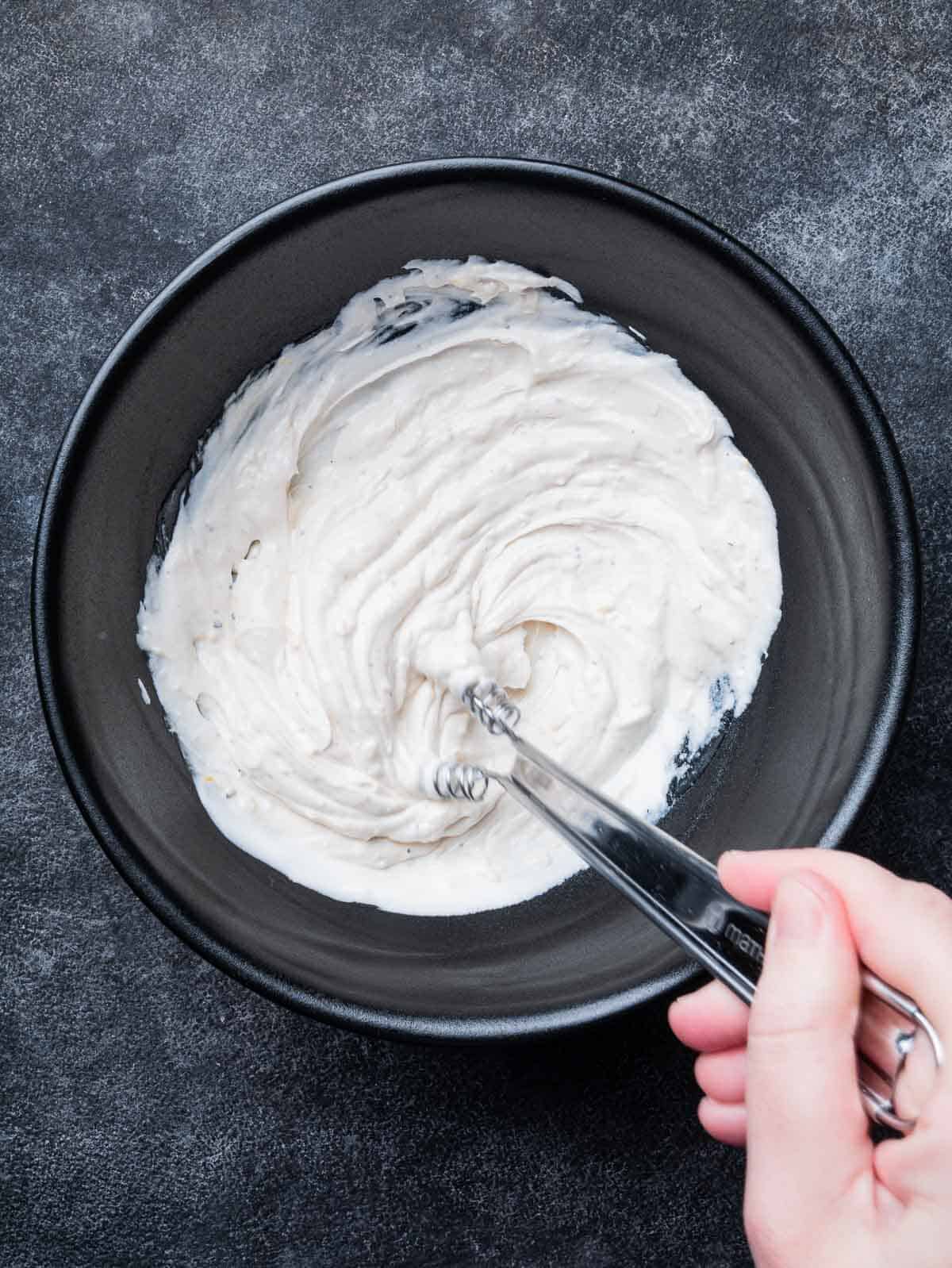 hand stirring yogurt tahini sauce in a black bowl.