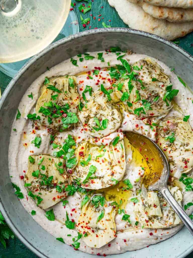 creamy white bean dip in a bowl topped with marinated artichoke hearts, parsley and chili flakes.