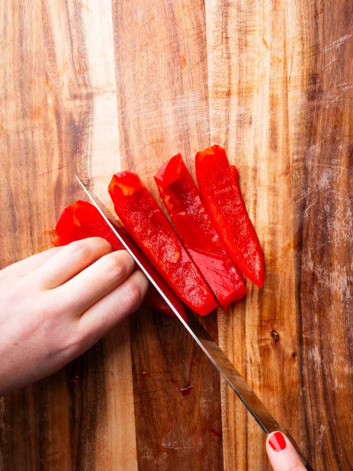Hands cutting a piece of red bell pepper into strips with a knife.