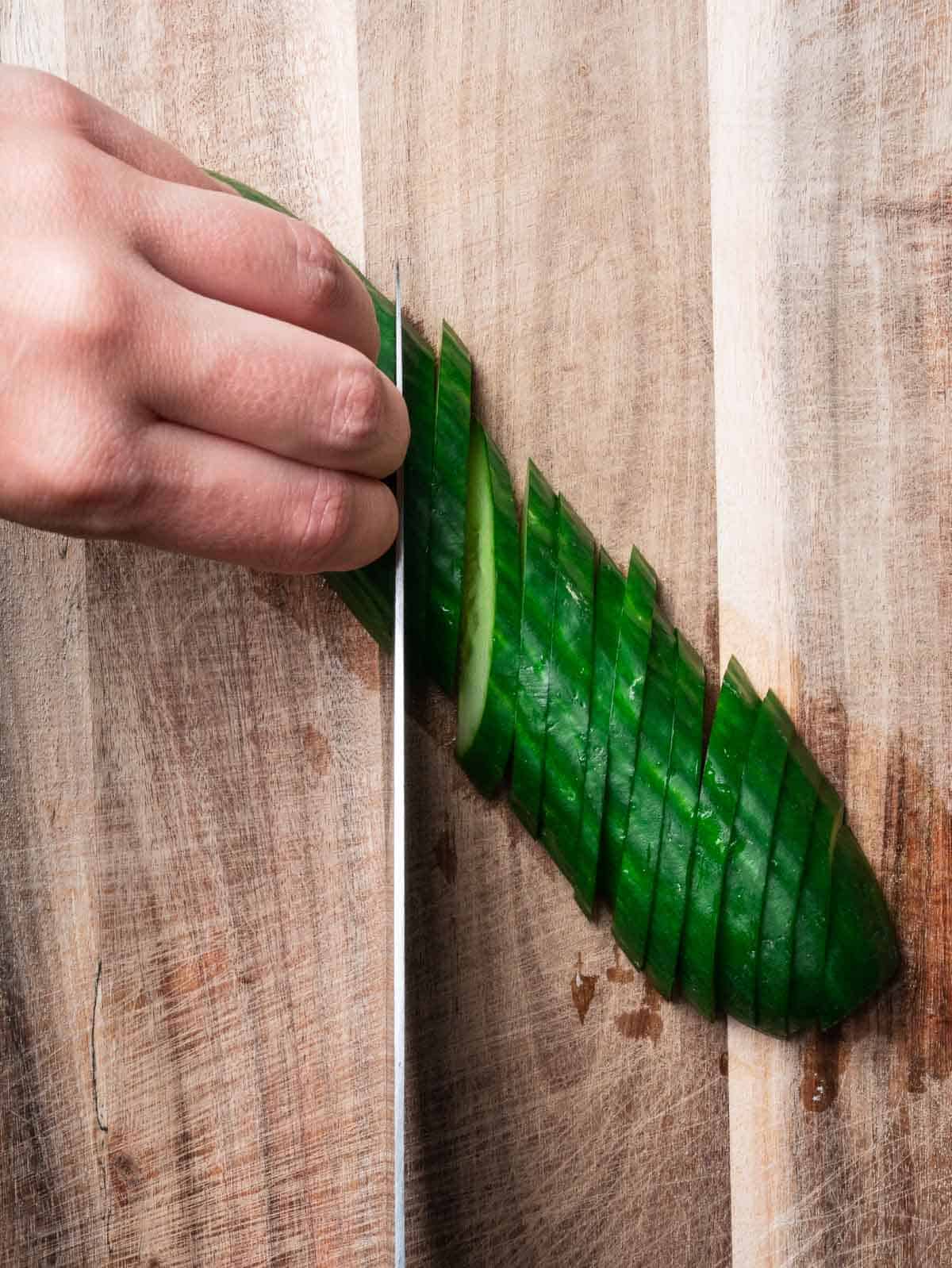 hand with a kitchen knife slicing cucumber diagonally.