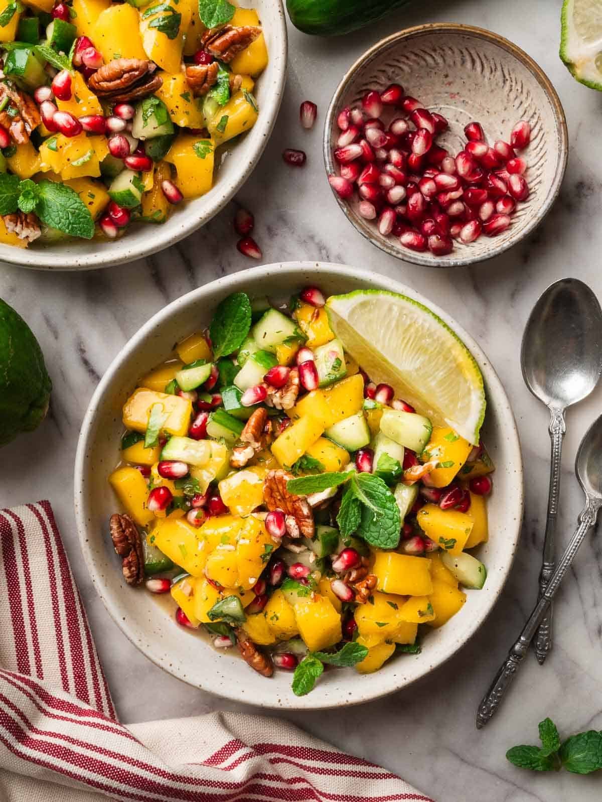 Two bowls of cucumber mango salad with pomegranate and spoons on the side.
