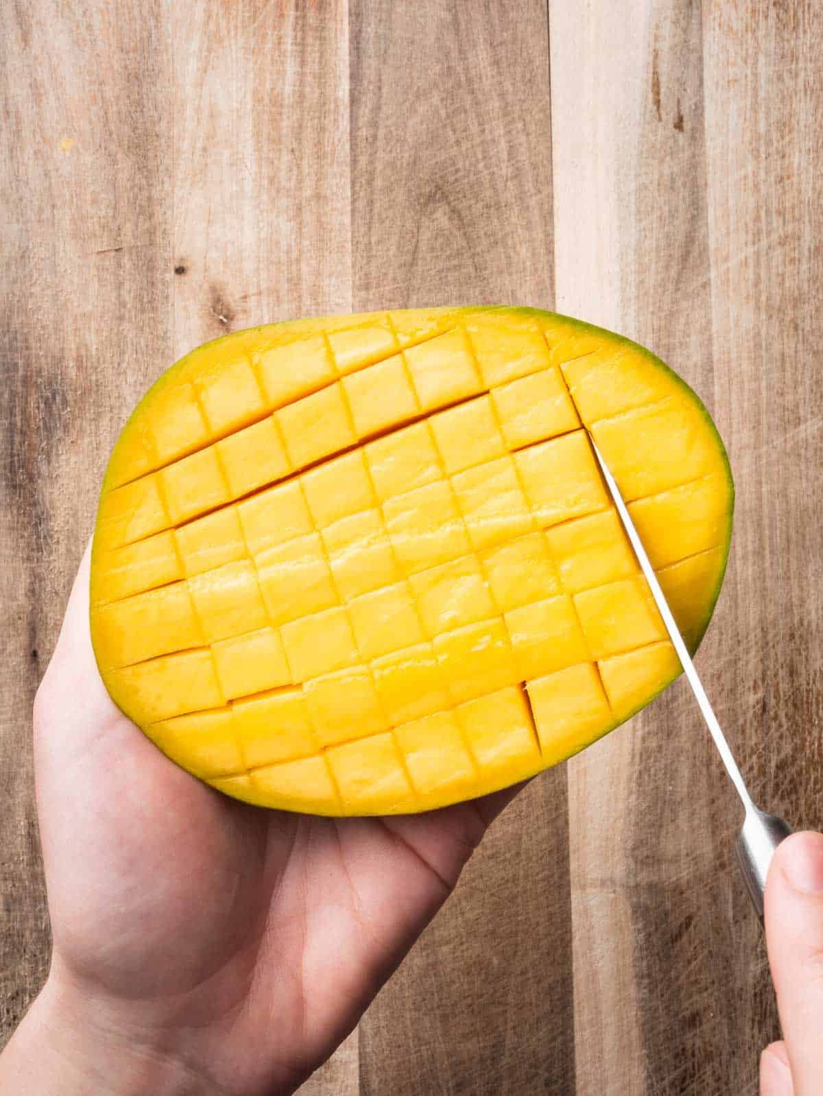 Hand with a knife scoring the flesh of a mango in a crosshatch pattern.