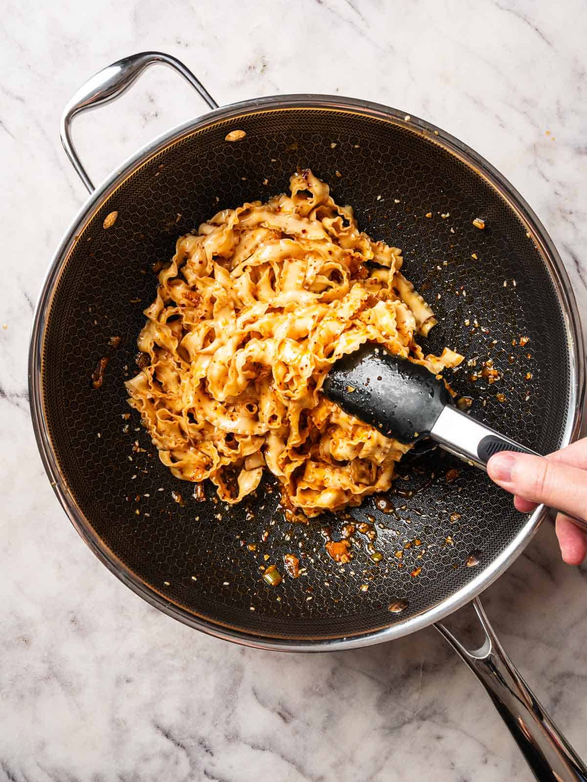 Kitchen tongs tossing noodles and garlic chili oil together.