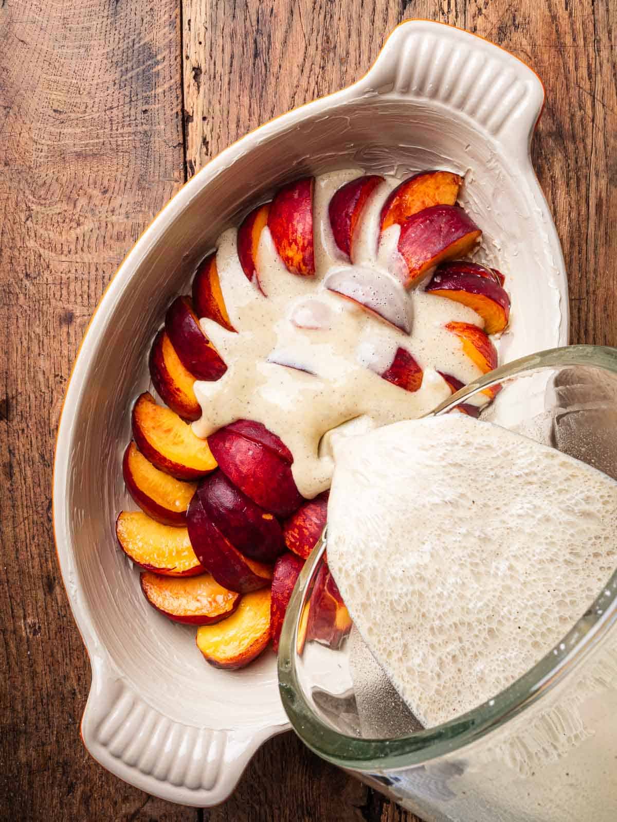 Clafoutis batter being poured on top of peaches in a baking dish.