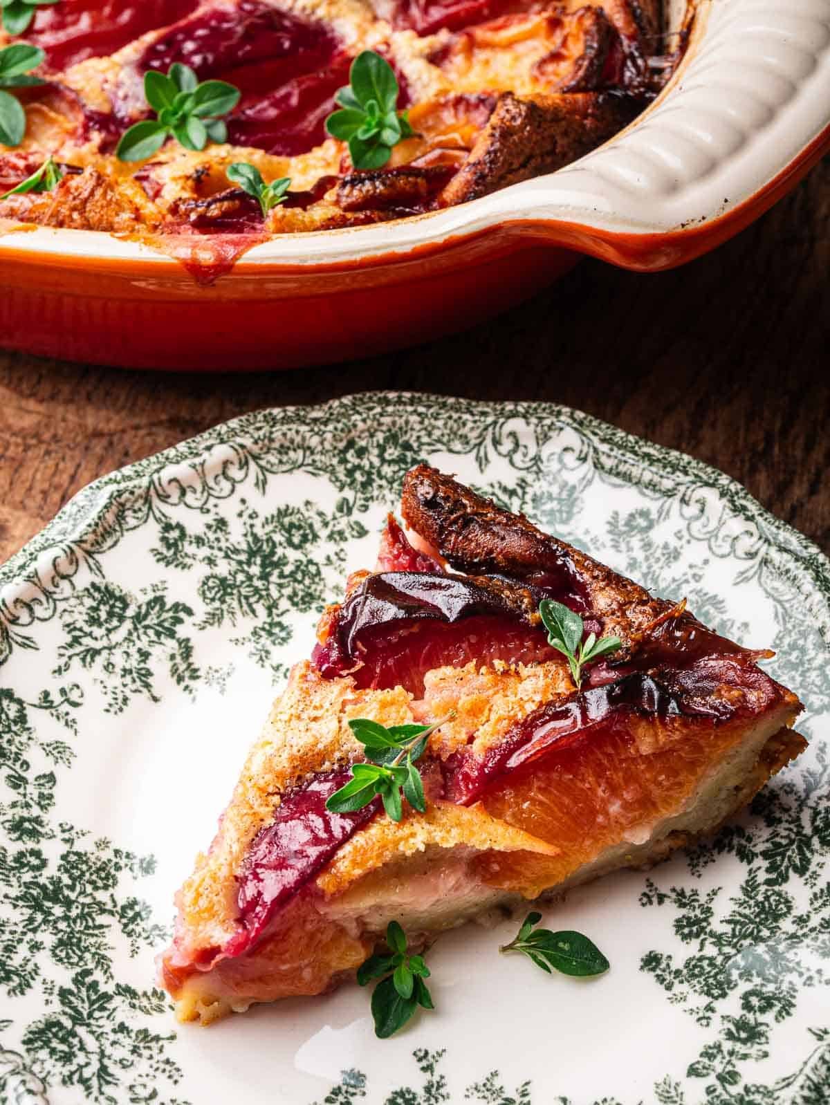 A slice of peach clafoutis on a vintage plate with the rest of the clafoutis in the background.