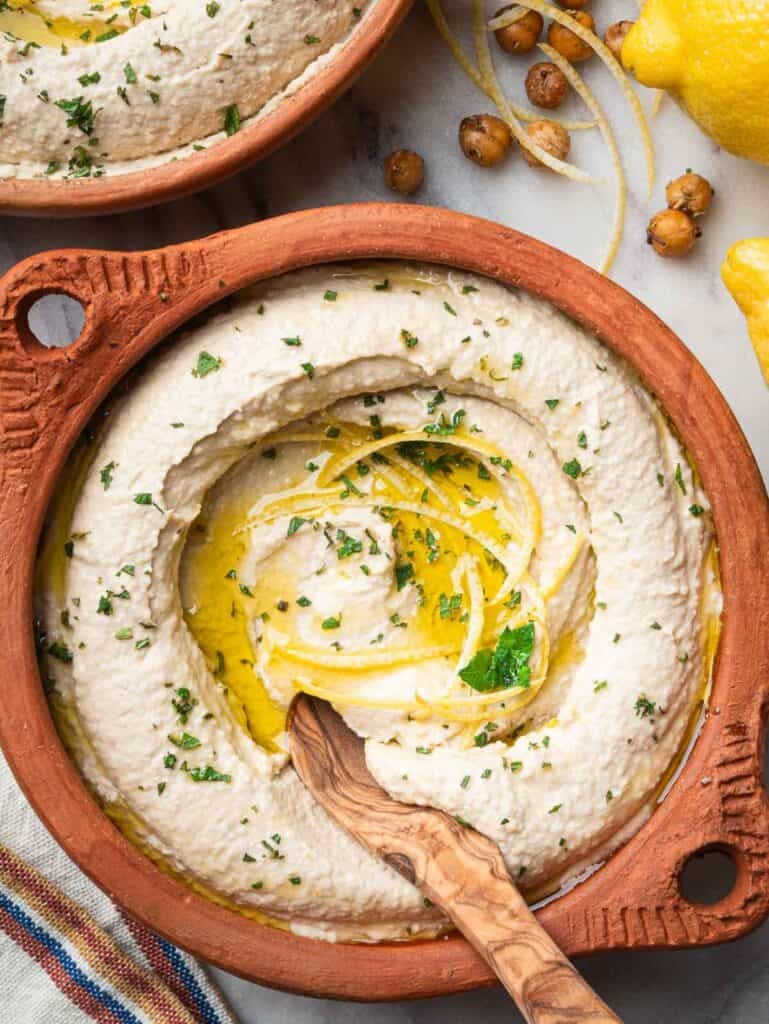 Lemon hummus topped with mint plated in a terracotta bowl with a wooden spoon.